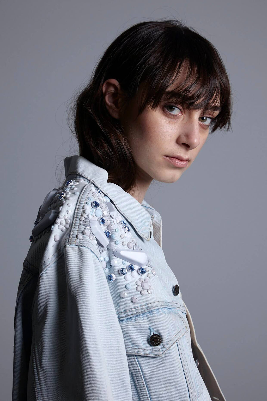 Close-up shot of the Pinball Jacket's hand-embellished details, showcasing the unique combination of clear triangular perspex domes, shimmering glass crystals, and matte white beads on light vintage denim.