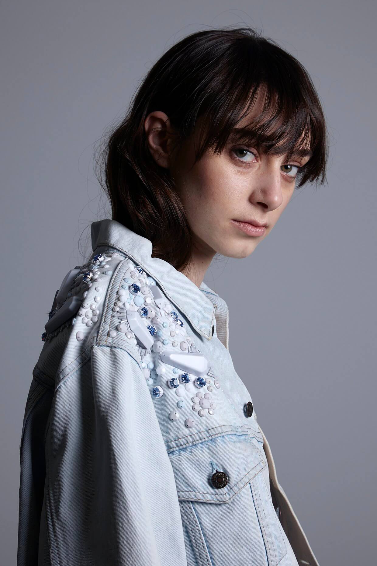 Close-up shot of the Pinball Jacket's hand-embellished details, showcasing the unique combination of clear triangular perspex domes, shimmering glass crystals, and matte white beads on light vintage denim.