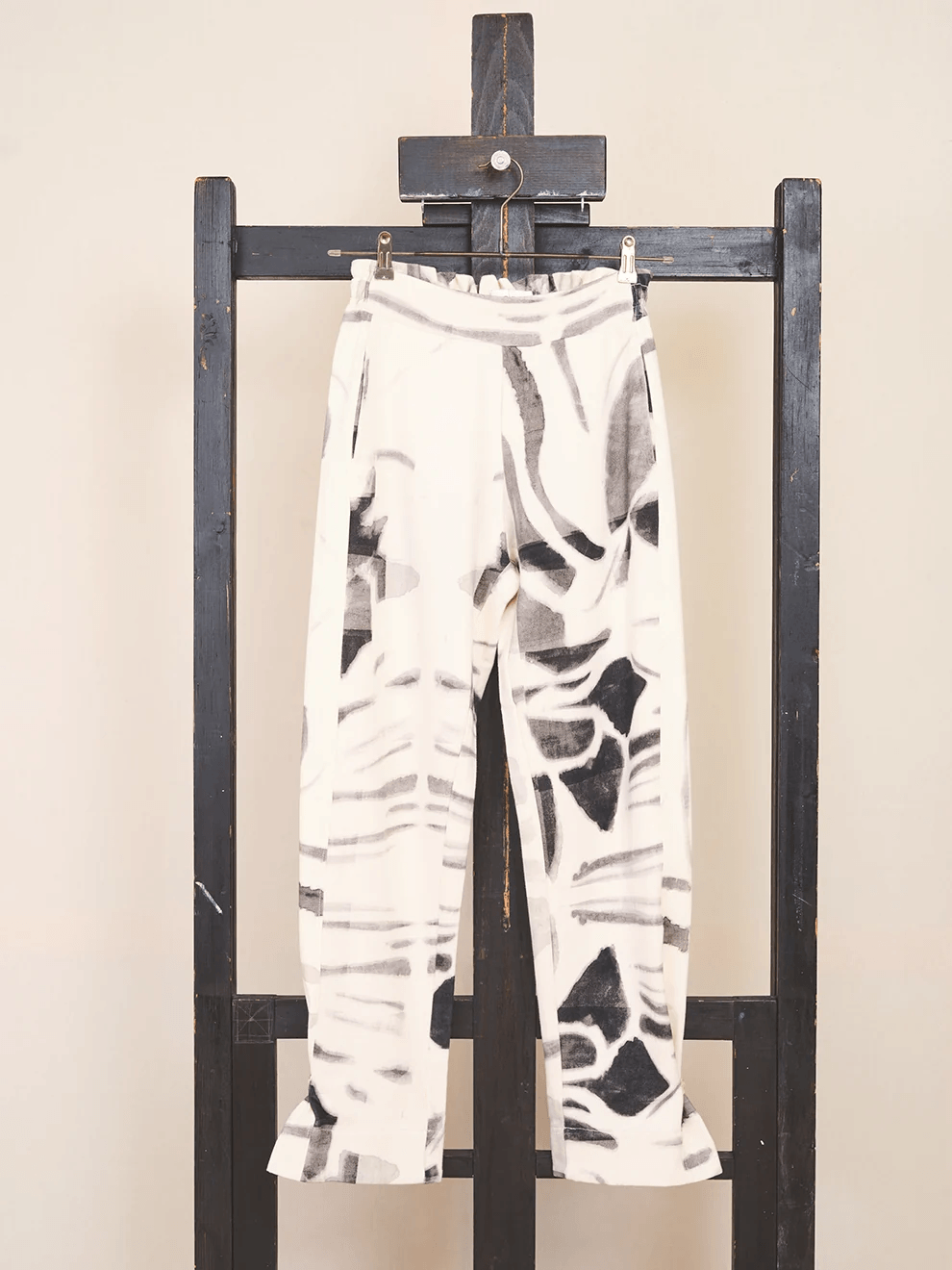 Front view of the limited capsule Pants 001 organic cotton trousers hanging from a minimalist wooden hanger showing the pure construction and art print pattern